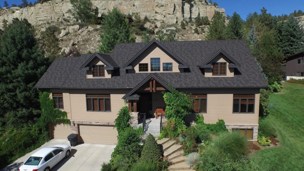 A large tan home with a charcoal grey stone coated steel roof, that sits at the base of a large rock