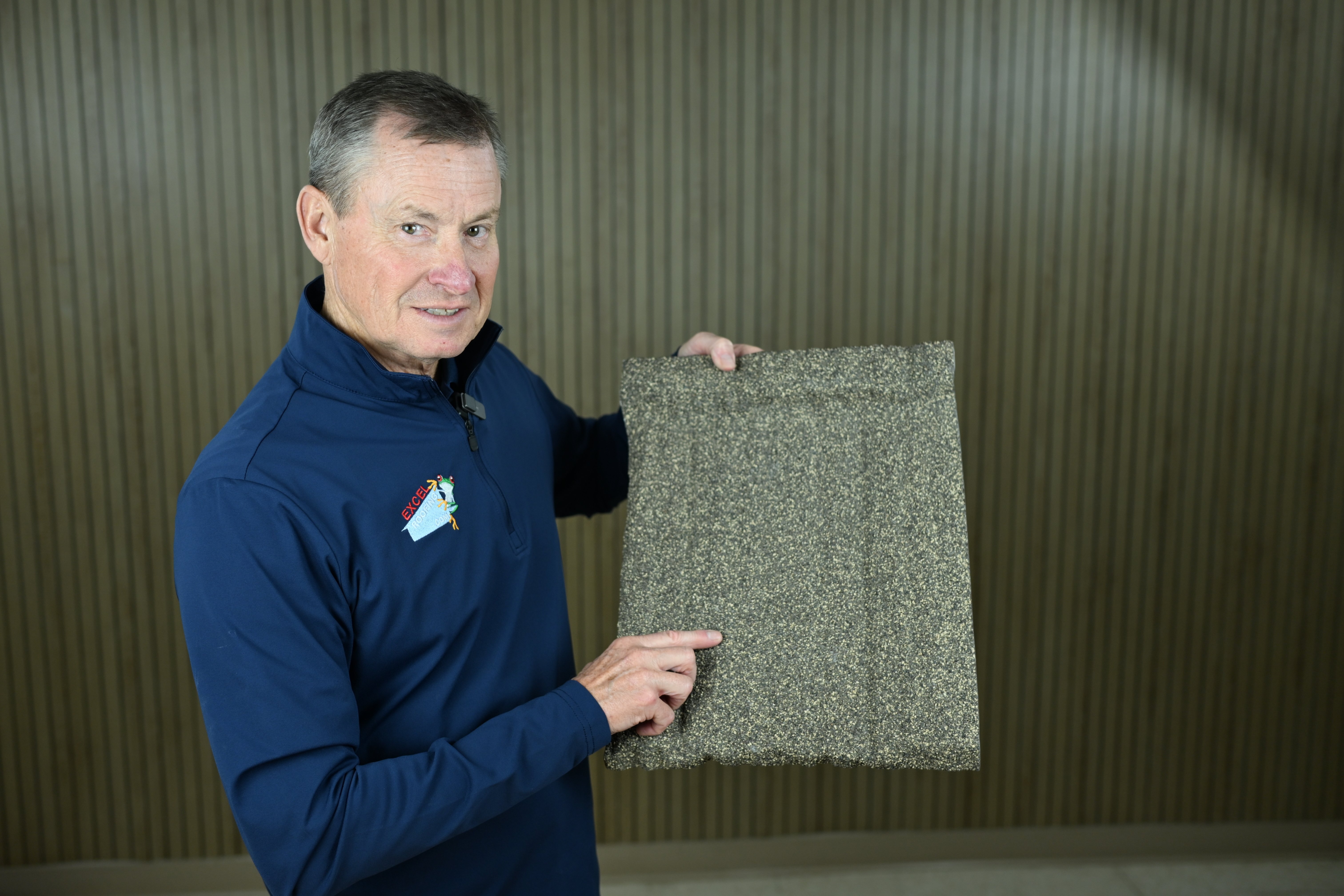 J Bretz of Excel Roofing in a dark blue long sleeve shirt holding a slate colored DECRA stone coated steel shingle