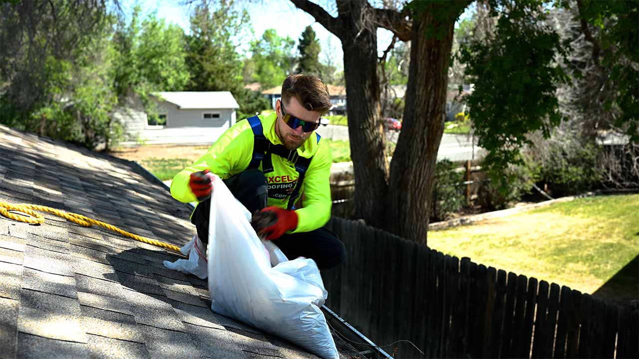 Zach McDonald Excel Roofing gutter expert, cleaning the gutters of a one story home. He is in a yellow high visibility Excel Roofing shirt and is secured with a safety harness and rope