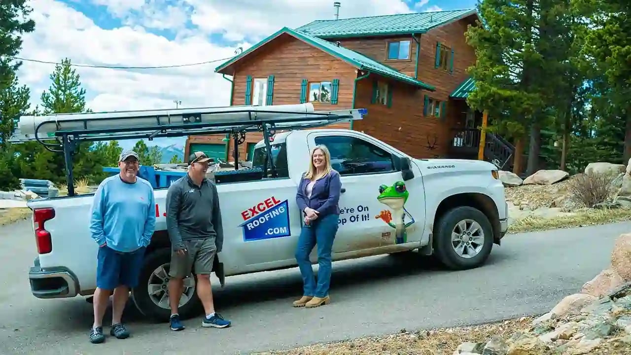 Happy customers standing in front of a white Excel Roofing truck, the truck is parked in front of their mountain home and Adam of Excel Roofing is standing with them