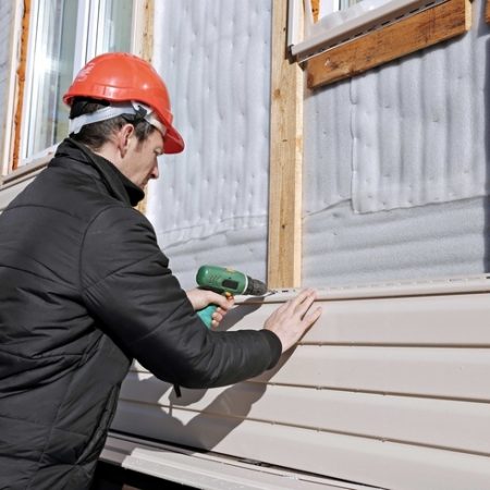 A kneeled worker wearing a red hard hat and a black jacket while installing tan siding on a house.