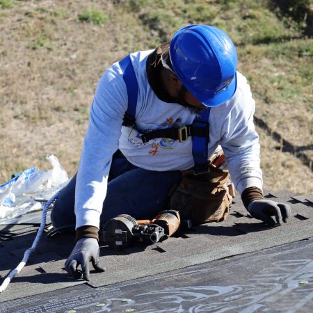 Man with a blue hard hat and harness sitting on roof, inspecting the work he has done.